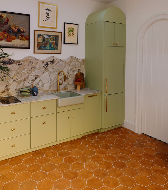 A kitchen features Zia Tile's 8x9 Hex + Fired Earth terra cotta floor, light green cabinets, a marble backsplash, gold faucet, framed art above the counter, and a tall green pantry beside an arched white door inspired by San Miguel de Allende.