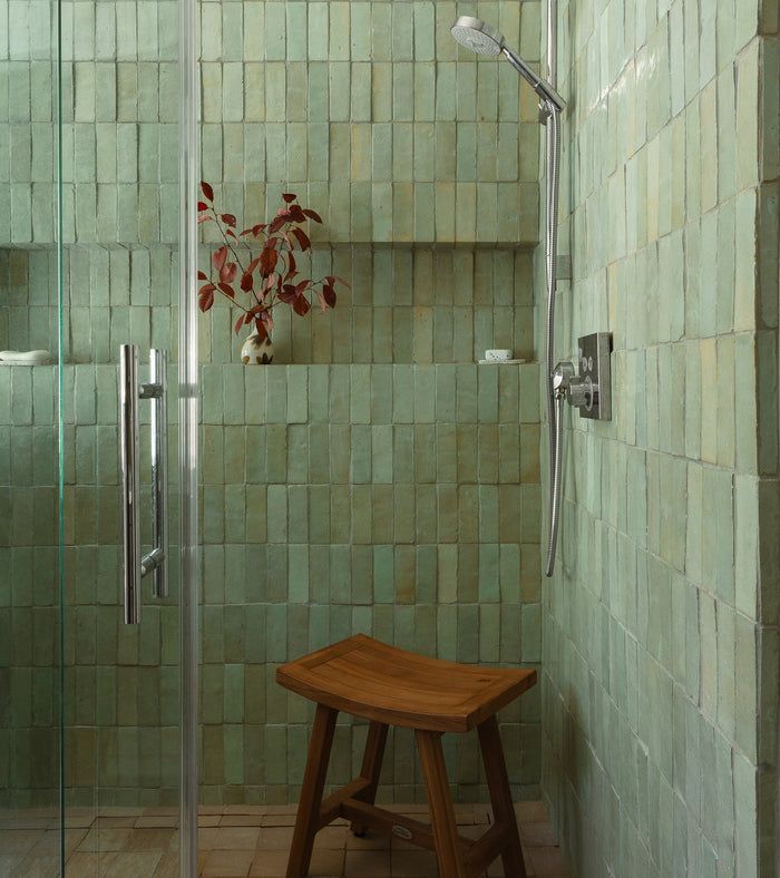 A shower featuring Absinthe 2x6 zellige tiles by Zia Tile on the walls, a modern showerhead, wooden stool, red-leafed plant on a built-in shelf, and a glass door.