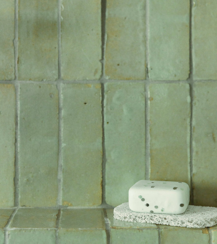 A white soap bar with dark specks sits on a natural sponge, set against a backdrop of Zia Tile’s Absinthe 2x6 green Moroccan tiles.