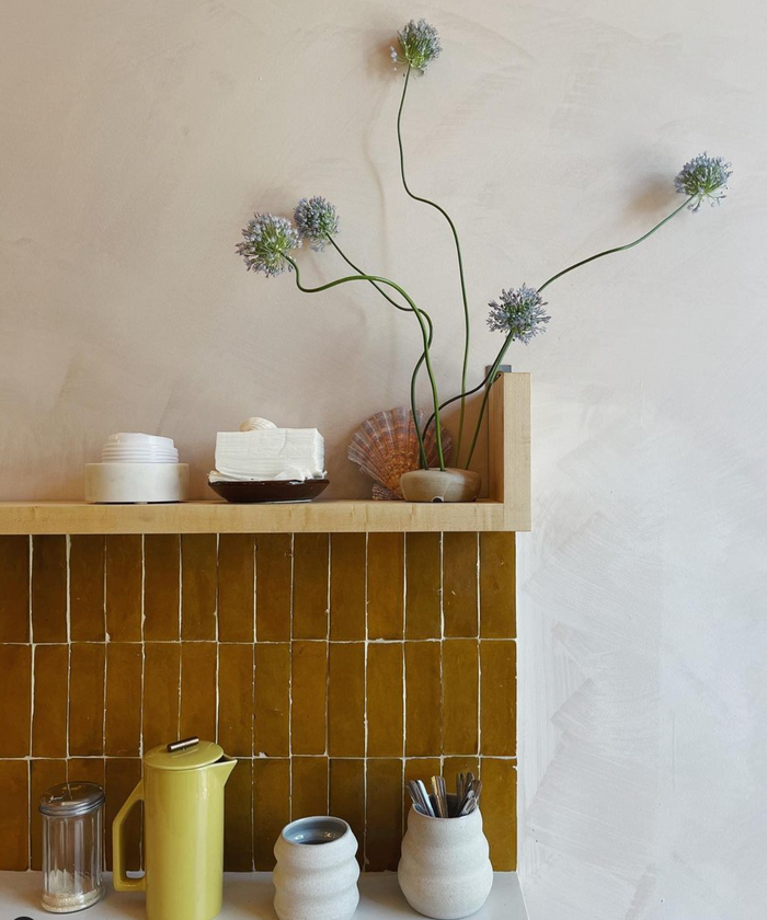 A shelf with a yellow Amber 2x6 Moroccan tile kitchen backsplash by Zia Tile displays ceramic dishes and a vase with four tall green stems topped with round purple flowers. Below, a yellow coffee pot, two mugs, utensils, and a glass jar sit on the counter.