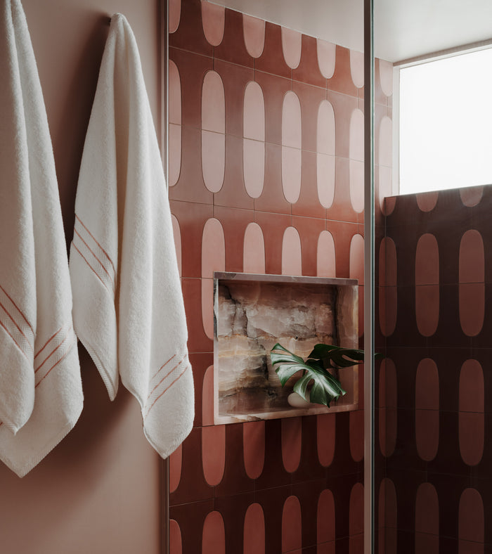 Two white towels hang on a pink wall beside a shower adorned with Zia Tile’s Arco Protea + Canyon handmade tile, featuring reddish-brown, oval-shaped patterns. A built-in niche holds a plant leaf and stones beneath a frosted window, showcasing the artisanal quality of Zia Tile craftsmanship.
