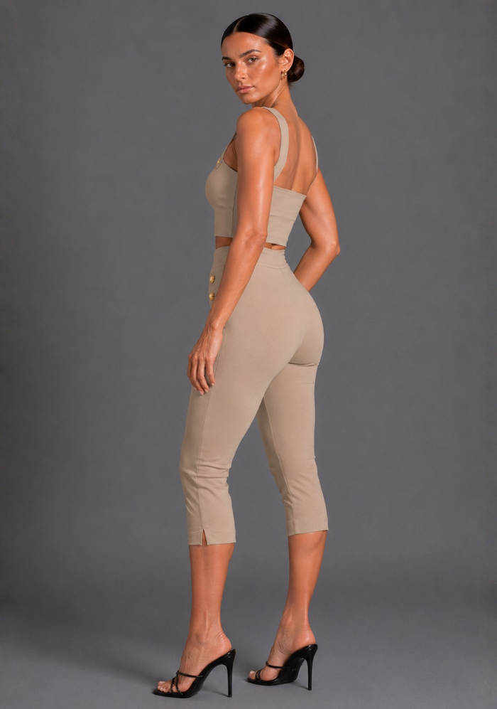 A woman modeling a beige two-piece activewear set consisting of a crop top and high-waisted capri pants, paired with black high-heeled sandals, against a solid gray studio background.