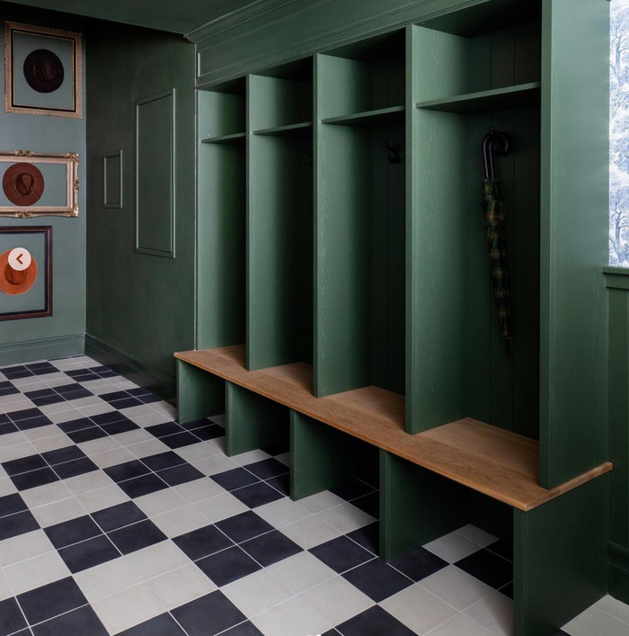 A green mudroom with four open cubbies featuring wooden benches. A plaid umbrella hangs in one cubby. The floor features Black 8x8 tile by vendor-unknown, mixed with white and gray squares. Framed hats hang on the left wall.