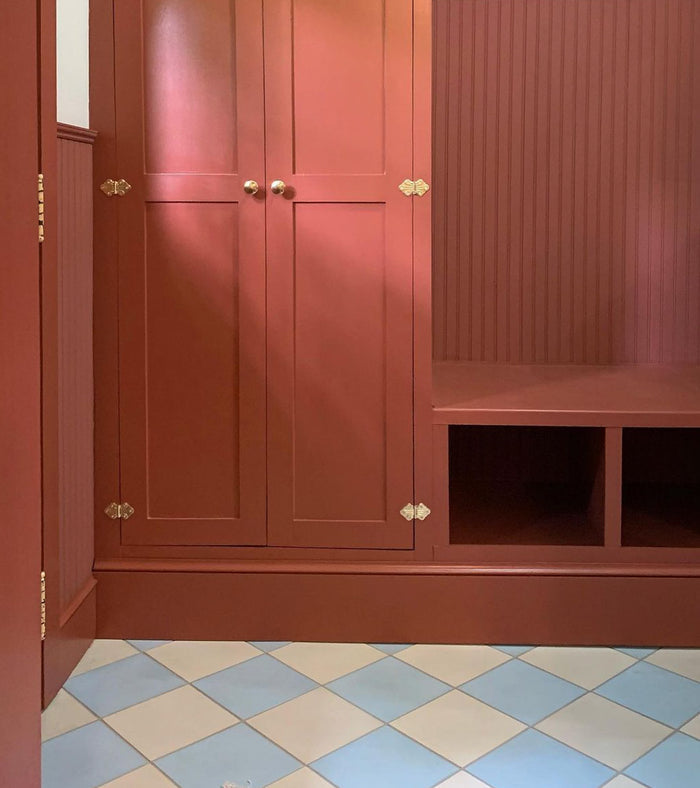 A built-in storage unit with paneled doors and open cubbies, all in muted reddish-brown, stands against a grooved wall. The floor is finished in vendor-unknown’s Bone 8x8 pale blue and white cement tile arranged in a diamond pattern.