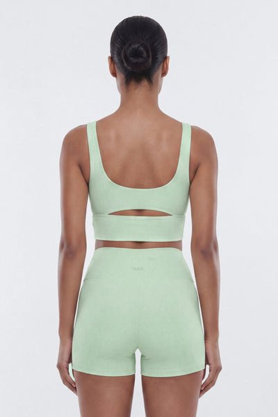 A woman shown from the back wearing a light mint green activewear set consisting of a sports bra with a horizontal cutout across the upper back and high-waisted shorts, against a plain light gray studio background.