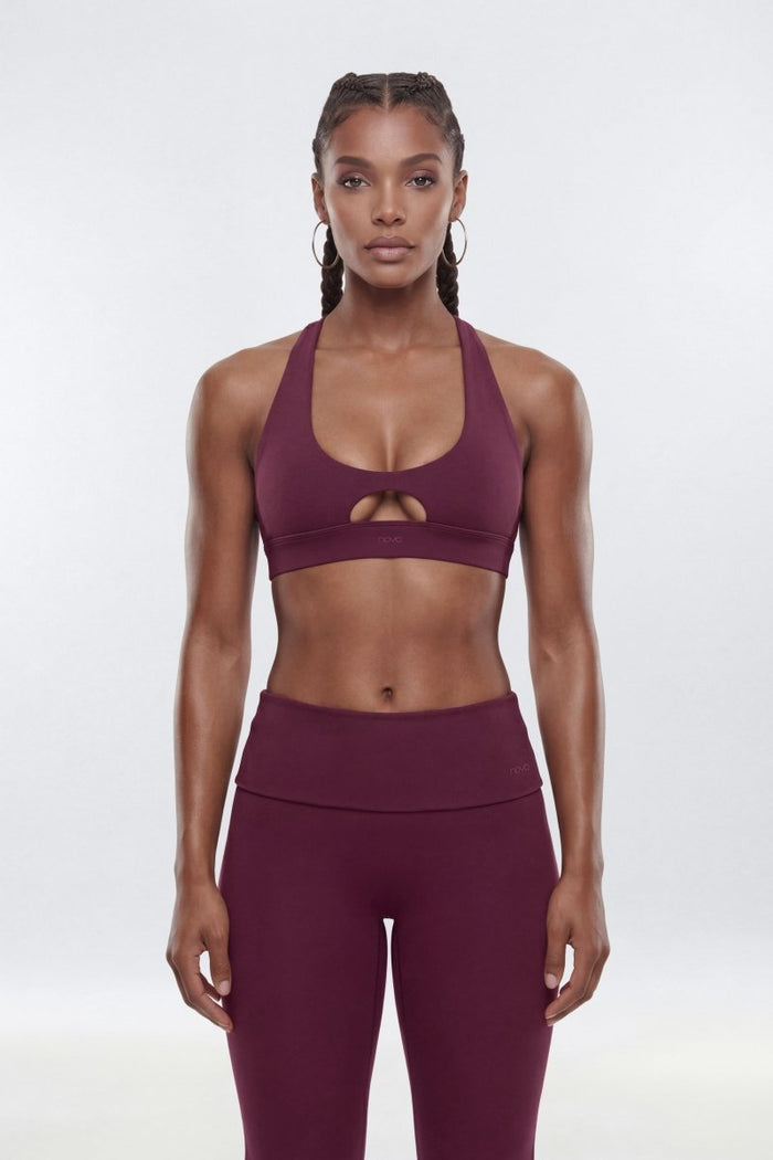 A woman modeling a matching athletic set consisting of a deep plum purple sports bra with a keyhole cutout and high-waisted leggings, shot against a seamless light gray studio background.