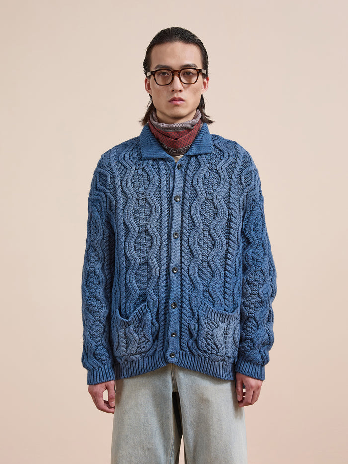 Someone wears Found's Cable Knit Buttoned Cardigan with front pockets, layered over a turtleneck and paired with light blue jeans and glasses, standing against a beige background.