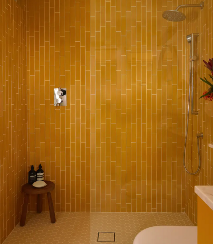 A modern shower features orange Cadmium 2x8 subway tiles by vendor-unknown, a rainfall showerhead with wall-mounted controls, and a wooden stool holding bottles. A partial view of a sink and a floral arrangement appear on the right.
