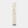 The Caldera 2x12 by Zia Tile is a tall, narrow rectangular slab of light terrazzo with small gray and beige stone fragments, displayed upright against a plain white background.