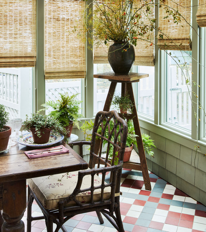 A sunroom with light green window frames, bamboo shades, potted plants on a wooden table and stand, a wicker chair with a floral cushion, and a Canyon 4x4 cement tile floor by vendor-unknown in patterned red, white, and blue.