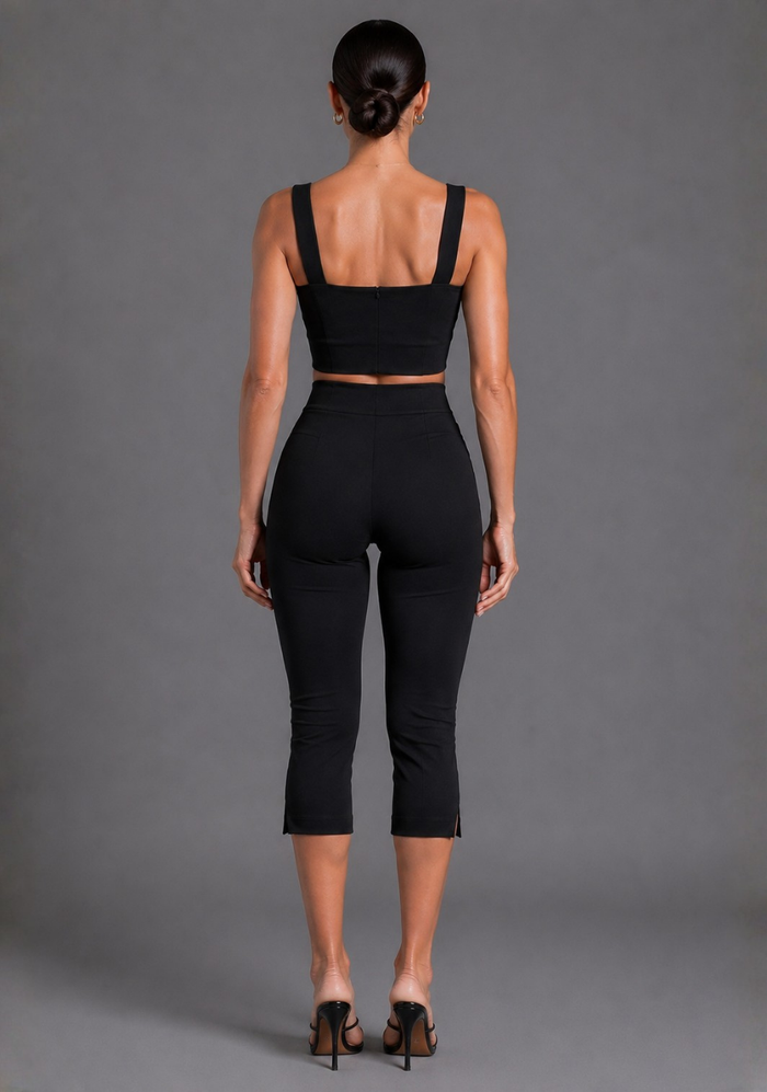 A woman viewed from the back wearing a black two-piece activewear set consisting of a cropped tank top and high-waisted capri pants, paired with black heels, against a solid gray studio background.