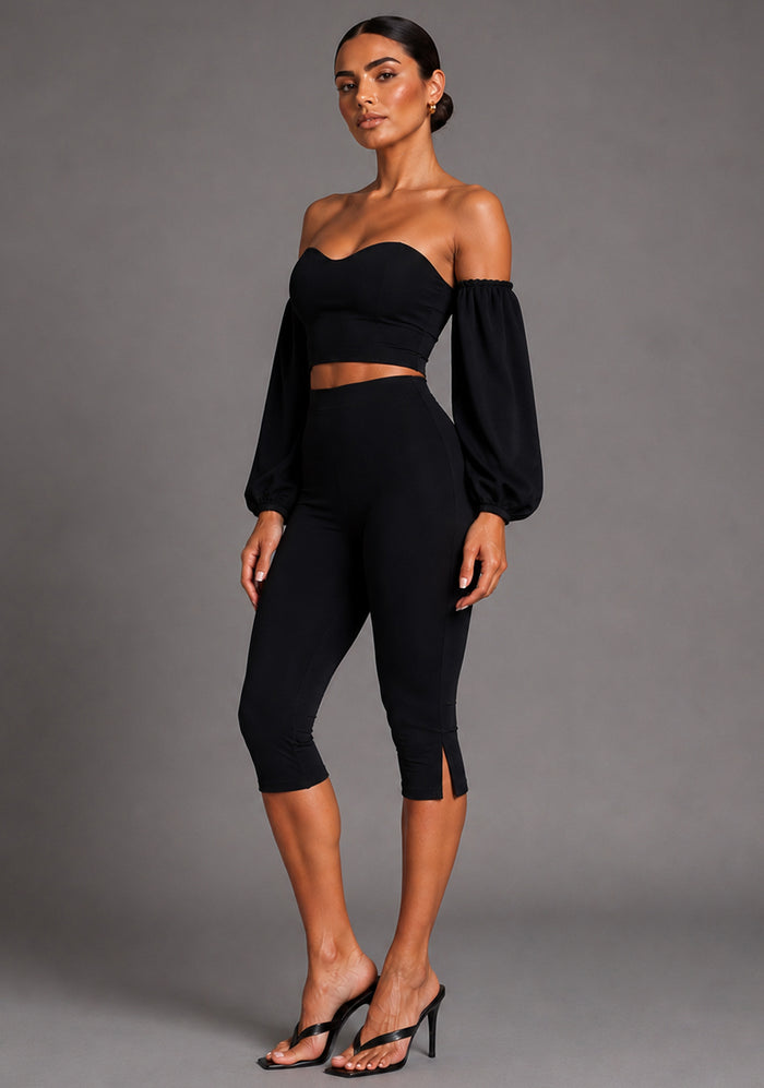 Side front image of a model wearing the VSPR Capri Cropped Pants in Black showcasing the sleek silhouette, flattering high-rise design and subtle side hem split detail against a grey studio backdrop