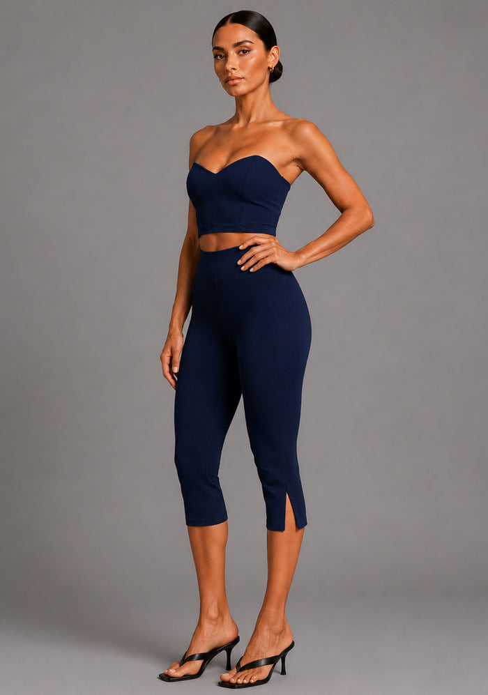 Side front image of a model wearing the VSPR Benedetta Strapless Navy Crop Top in navy, showcasing the sleek strapless design and figure-enhancing fit. The design paired with Capri Cropped Navy Trousers, against a grey studio backdrop.