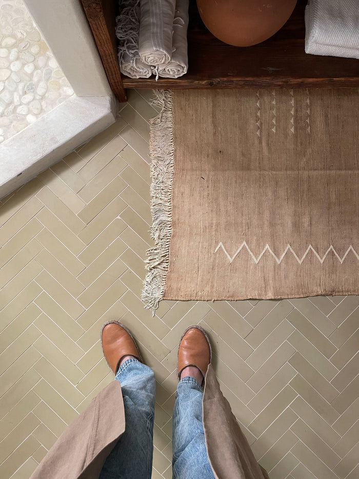 A person in tan shoes, blue jeans, and a beige coat stands on a herringbone floor near a geometric woven rug; rolled towels and pottery are on a shelf by vendor-unknown's Clay 2x8 cement tile kitchen backsplash.
