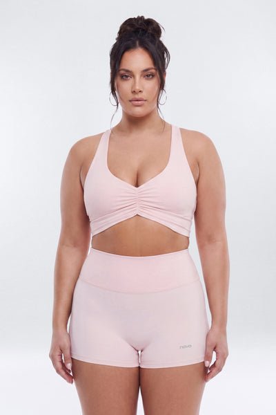 A woman modeling a light pink two-piece activewear set consisting of a sports bra with a front cutout and ruched detail, paired with matching high-waisted shorts, against a plain light gray background.