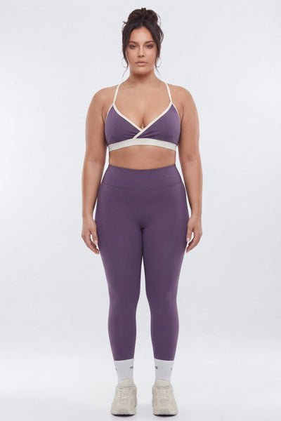 A woman modeling a matching purple activewear set consisting of a sports bra with white contrast trim and high-waisted leggings, paired with white socks and beige sneakers, against a light gray studio background.