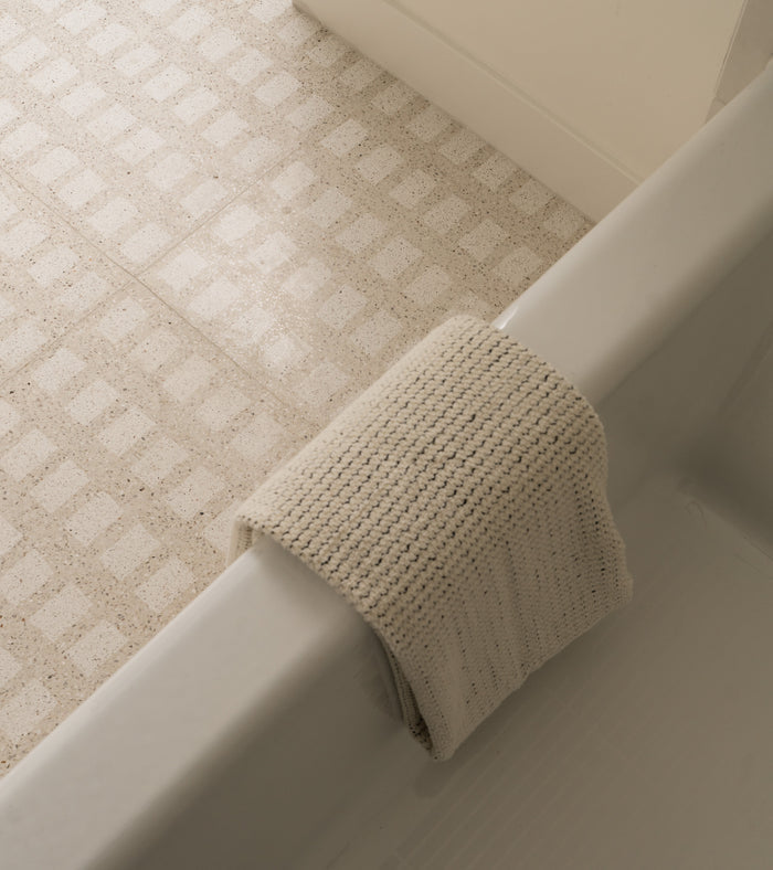 A knitted beige washcloth is draped over the edge of a white bathtub, with sleek Constellation Bone + White 24x24 terrazzo cement tile flooring by Zia Tile visible in the background.