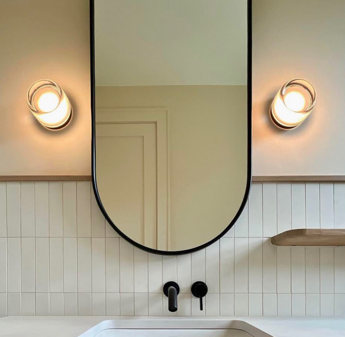 A modern bathroom features a large oval mirror with black trim, round wall lights, and a white sink with black fixtures. The backsplash uses Cotton 2x8 tiles by vendor-unknown, complemented by a wooden shelf on the right.
