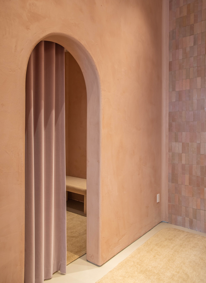 A soft pink arched doorway with a matching velvet curtain opens to a small bench; warm, muted pink walls are accented by Zia Tile’s Desert Bloom 2x6 tiles, while a beige carpet covers the floor.