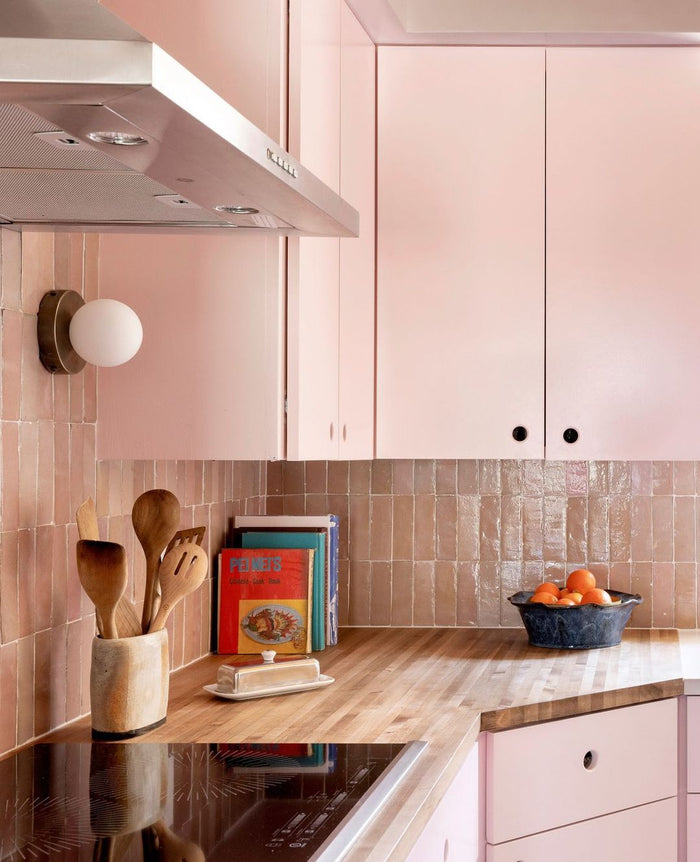 A modern kitchen corner with blush pink cabinets, a light wood countertop, a pink tile backsplash, and a built-in black cooktop. A bowl of oranges, a cup with wooden utensils, and a stack of cookbooks sit on the counter.