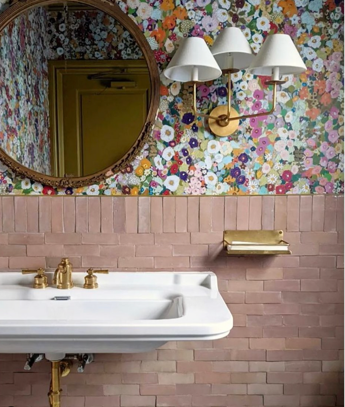 A bathroom features a white sink, brass fixtures, a gold wall-mounted soap holder, Desert Bloom 2x6 tiles by Zia Tile on the lower wall, colorful floral wallpaper above, a round mirror, and a brass double light fixture.