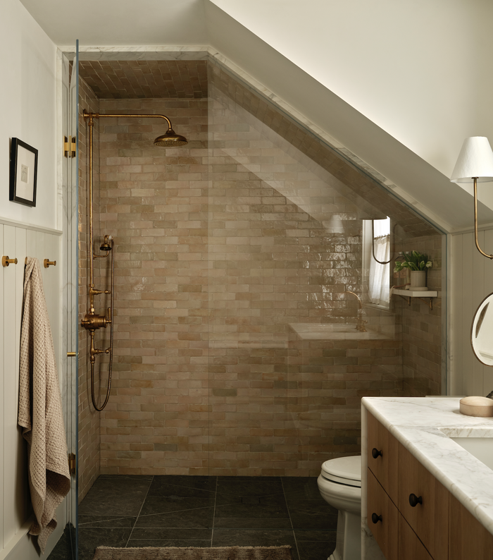 A bathroom with a sloped ceiling features a walk-in shower with beige and Zia Tile Glazed Earth 2x6 tiles and gold fixtures, plus a towel on a hook, marble-topped vanity, toilet, and soft lighting.