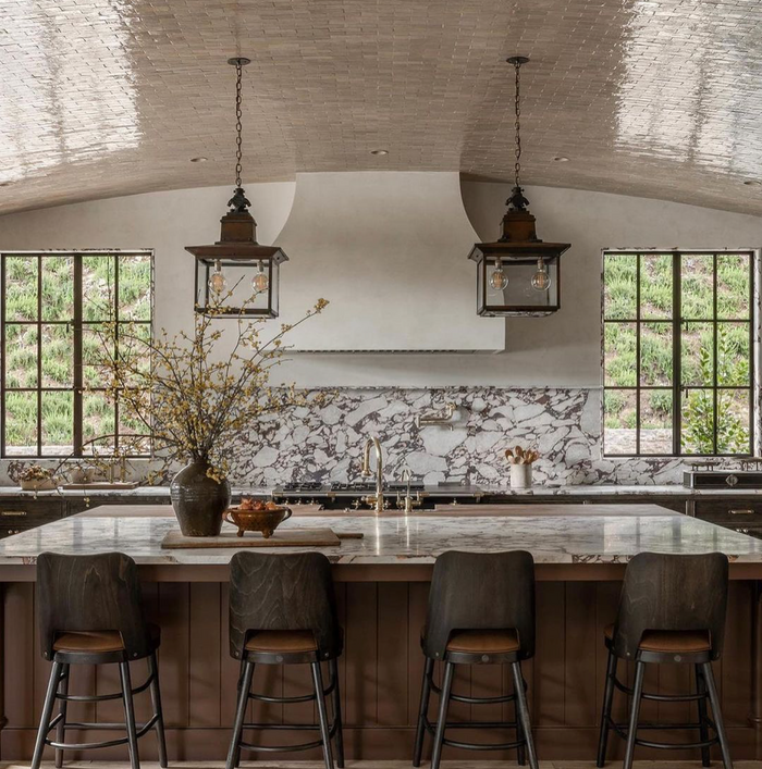 A stylish kitchen with a large island, four dark stools, marble countertop, pendant lights, a vase with branches, and wide windows. The backsplash features dramatic marble patterns and Glazed Earth 2x6 tiles by Zia Tile.