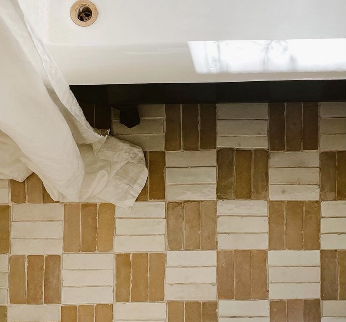 A white bathtub with a drain sits beside a floor of Glazed Earth 2x6 tiles from Zia Tile, arranged in a geometric tan and cream pattern. A white curtain draped over the tub adds softness to the Moroccan-inspired design.