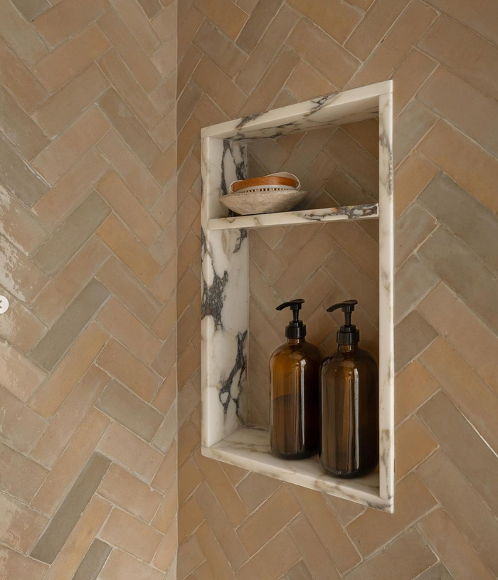 A marble shower niche holds two brown pump bottles below and a small bowl above, set against tan Zia Tile Glazed Earth 2x6 tiles in a herringbone pattern.