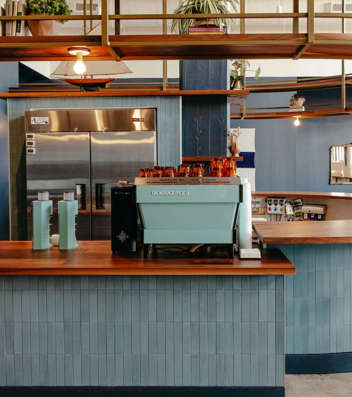 A modern coffee shop counter features a teal La Marzocco espresso machine, wooden countertop, metallic fridge, shelves with plants and cups, and a vendor-unknown Hyannis 2x8 subway tile backsplash in blue tones.