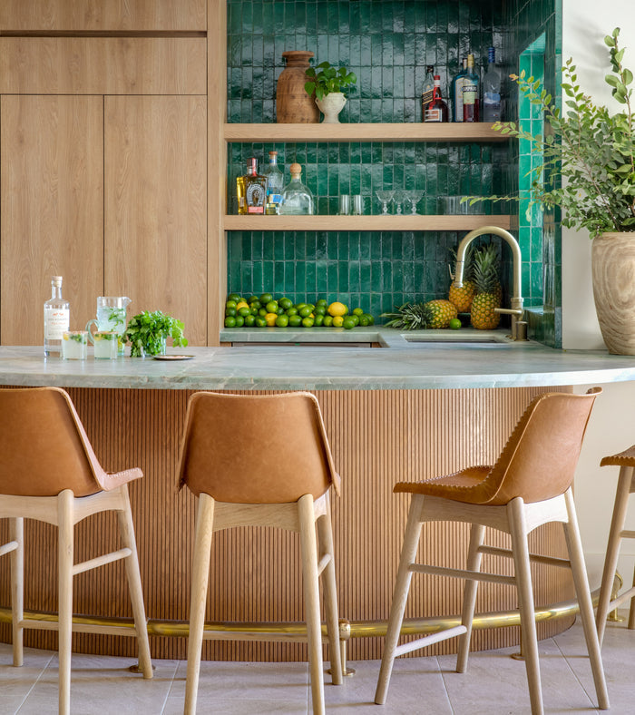A modern kitchen bar features tan leather stools, a marble countertop, wood cabinetry, open shelves with décor, fresh limes and pineapples, and a striking backsplash of Jade 2x6 tiles by Zia Tile behind the sink and vase with greenery.