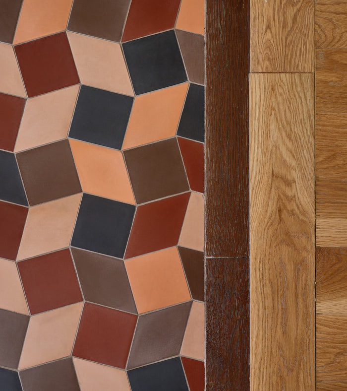 The Madrid Set One by Zia Tile features a bold 3D cube pattern in red, orange, brown, and black beside wooden floorboards in contrasting light and dark brown hues.