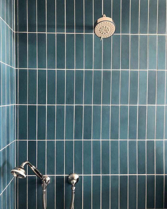 A shower clad in glossy Midnight 2x8 subway tile by vendor-unknown with white grout, featuring a round wall-mounted showerhead above and a lower handheld showerhead with hose.