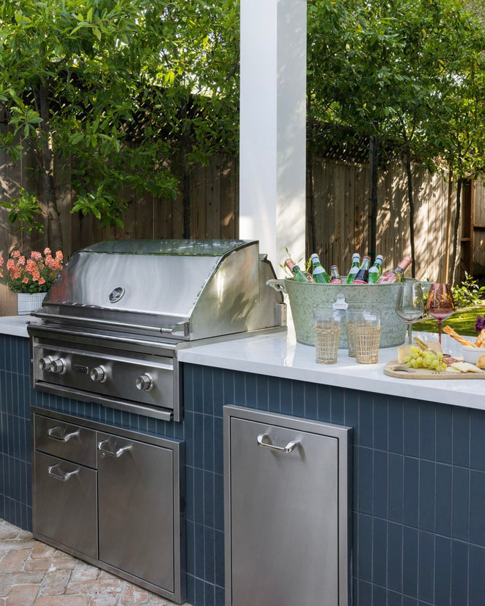 An outdoor kitchen with a stainless steel grill, storage cabinets, drinks, and grapes on a Midnight 2x8 blue cement tile counter from vendor-unknown that matches the backsplash, surrounded by lush greenery and flowers.
