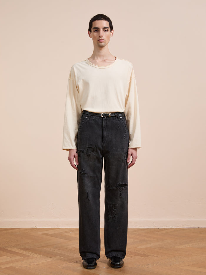 A person stands indoors against a plain beige wall, wearing a loose off-white long-sleeve shirt, FOUND Painter Repaired Jeans with a black belt, and black shoes.