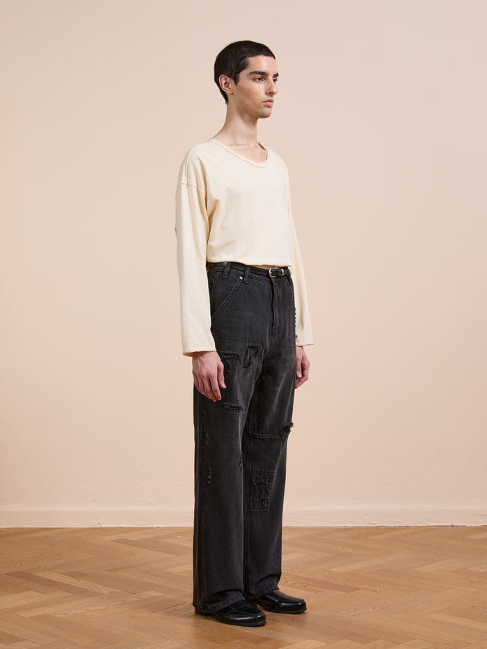 A person stands indoors on a wooden floor, facing left against a beige wall, wearing a cream long-sleeve shirt, black shoes, and FOUND Painter Repaired Jeans—a vintage wide-leg denim style.