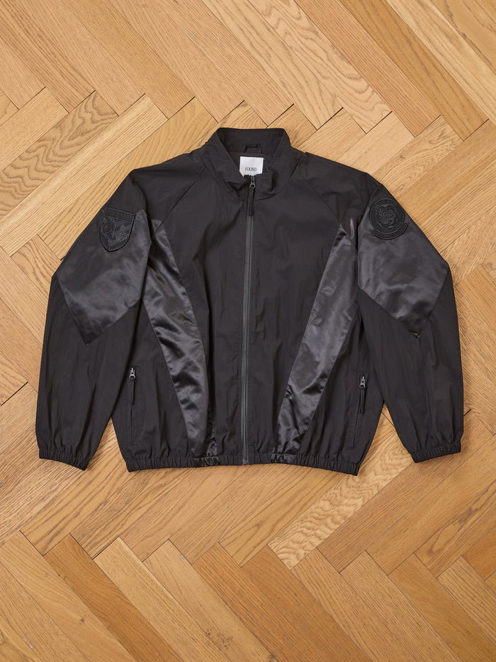 The FOUND Pine and Rose Jacket, a vintage sportswear-inspired black zip-up with satin panels and embroidered shoulder patches, is displayed flat on a light wooden herringbone floor.