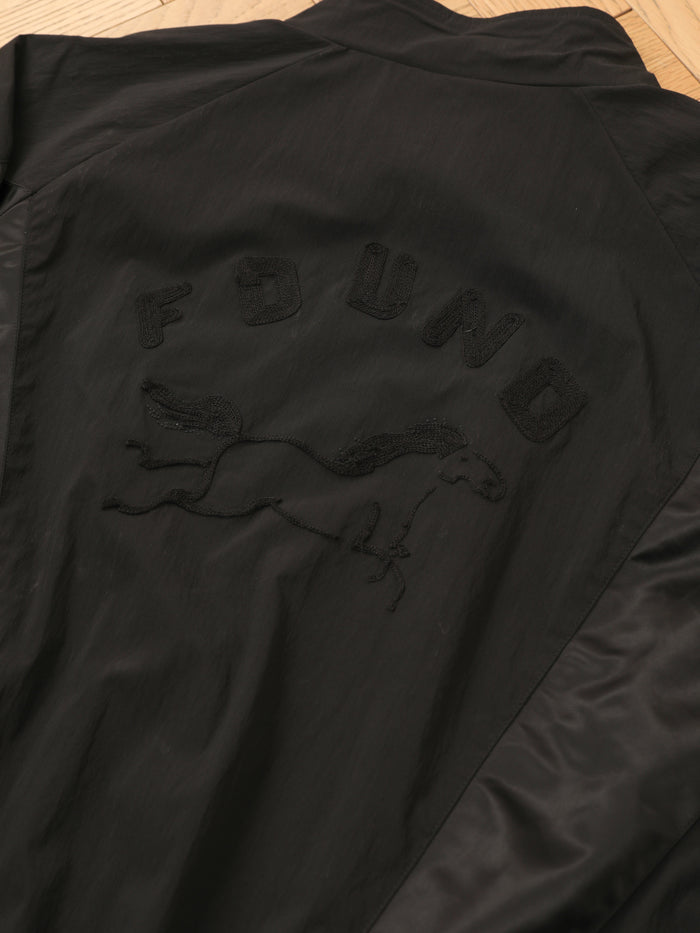 The FOUND Pine and Rose Jacket, a black vintage sportswear track jacket, is laid flat on wood with "FOUND" and a horse graphic embroidered on the back.