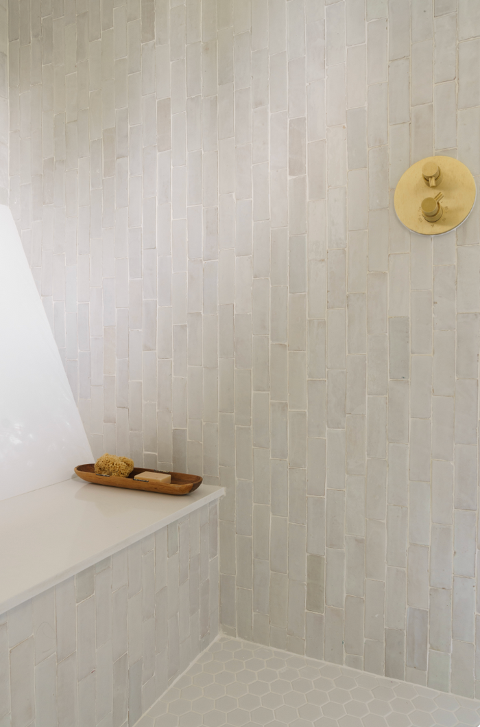 Minimalist shower featuring Zia Tile’s Pure White 2x6 tiles set vertically, a gold wall-mounted fixture, and a built-in bench with a wooden tray holding sponge and soap. Pale hexagonal tiles cover the floor.