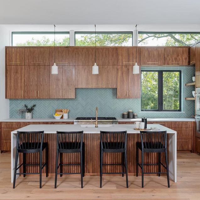 Modern kitchen with wood cabinets, large windows, and a white island with vertical wood accents. The Sage 2x8 tiles by vendor-unknown create a pale blue and sage green backsplash. Black bar stools, pendant lights, and light wood floors complete the look.