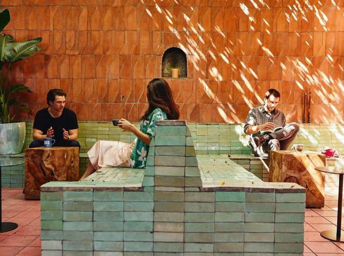Three people sit on wooden stools in a sunlit courtyard, where tree shadows move across peach and green Zia Tile Tidepool 2x6 tiles—creating a warm, relaxed vibe reminiscent of a kitchen backsplash as two chat and one reads.