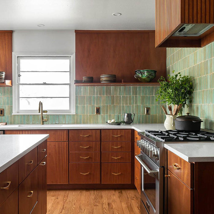 A modern kitchen with wood cabinets, white countertops, and a gas stove has a large window and a green backsplash of Tidepool 2x6 tiles by Zia Tile. Open shelves display dishes and a green bowl; a potted plant sits by the stove on light wood floors.