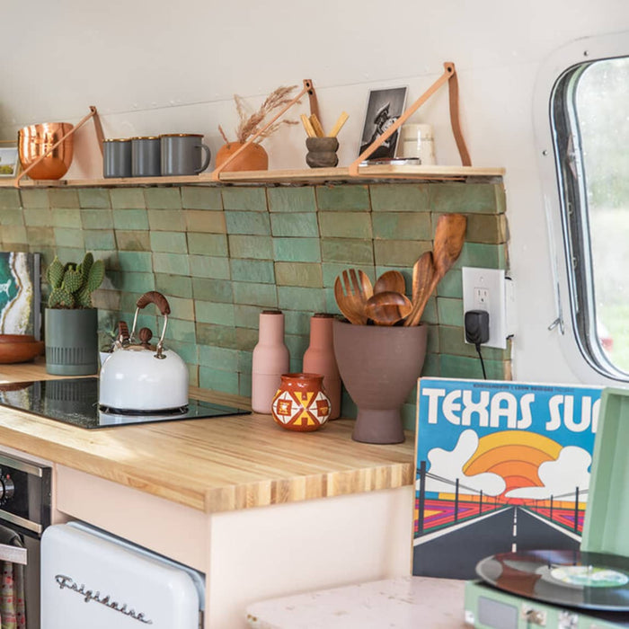 A cozy kitchen features a Zia Tile Tidepool 2x6 green backsplash, wooden countertop, ceramic and wooden utensils, a stove kettle, potted plants, and a colorful Texas Sun poster.