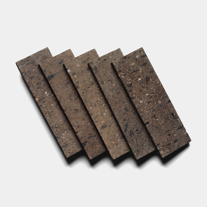 Five Zia Tile Volcan 4x12 rectangular terrazzo tiles, featuring a brown base with dark and light specks inspired by Mexican Cantera stone, are arranged diagonally and overlapped on a white background.